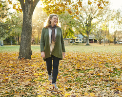 A woman walks through fallen leaves in a park.