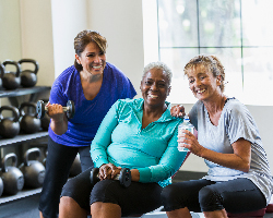 Three smiling women on a weight bench at a gym.