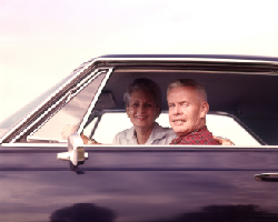 A smiling older couple seen through the window of an older model car.