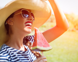 A smiling woman in a hat and sunglasses holding a slice of watermelon on a stick.