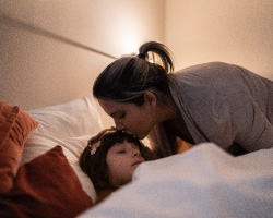 A woman kisses the forehead of a girl tucked into bed.